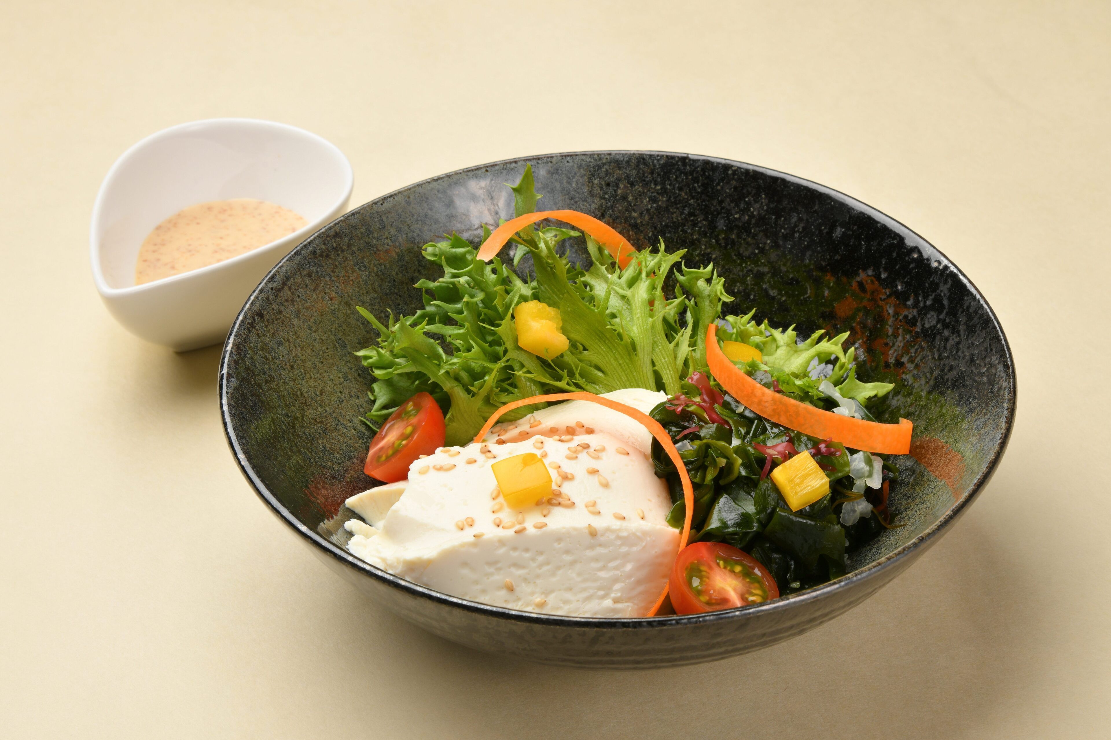 Ensalada con diversos vegetales y un trozo de tofu, acompañada de un recipiente pequeño con condimentos.