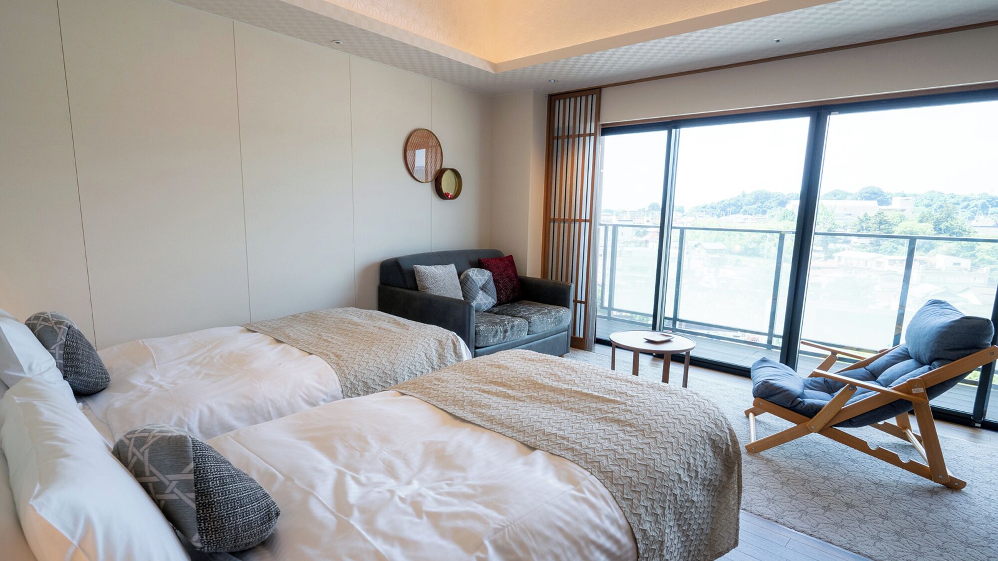 A hotel room with two beds, a sofa, a chair, a small table, and large windows offering a view.