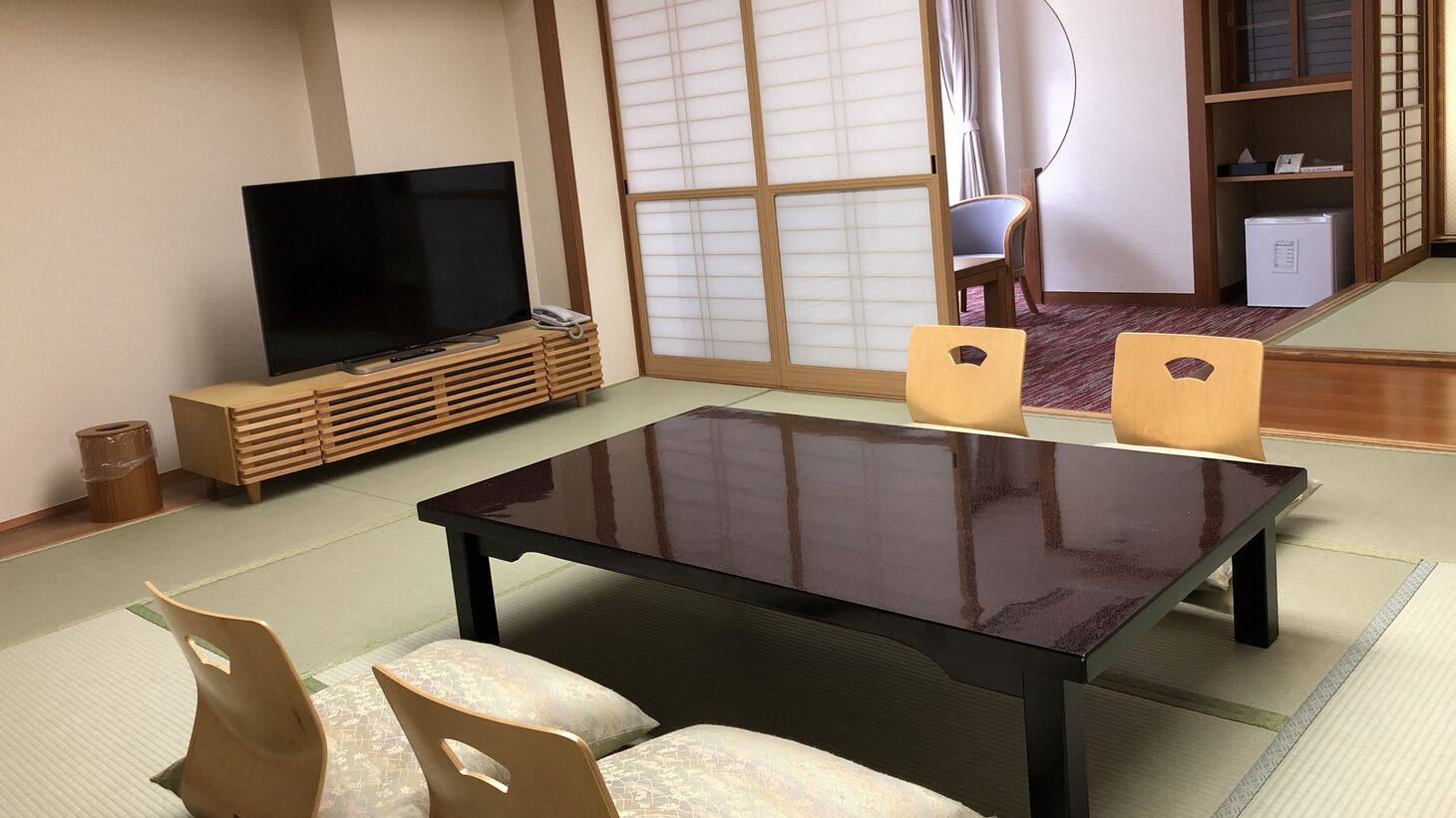 A traditional Japanese-style room with tatami flooring, a low table, two wooden chairs, a television, and a sliding door with shoji screens.