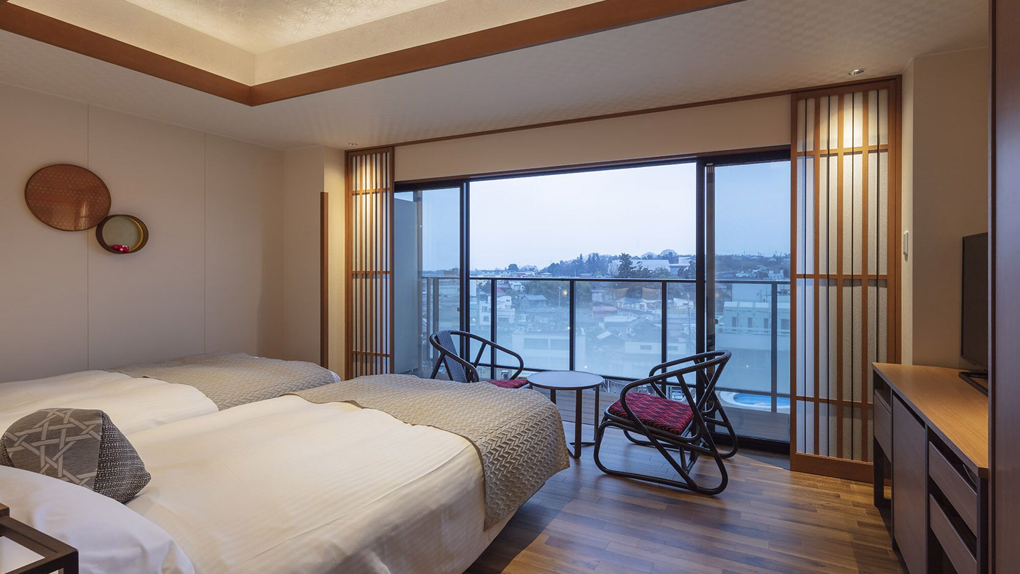 A hotel room with two beds, a small table, a chair, a TV, and a large window with a city view.