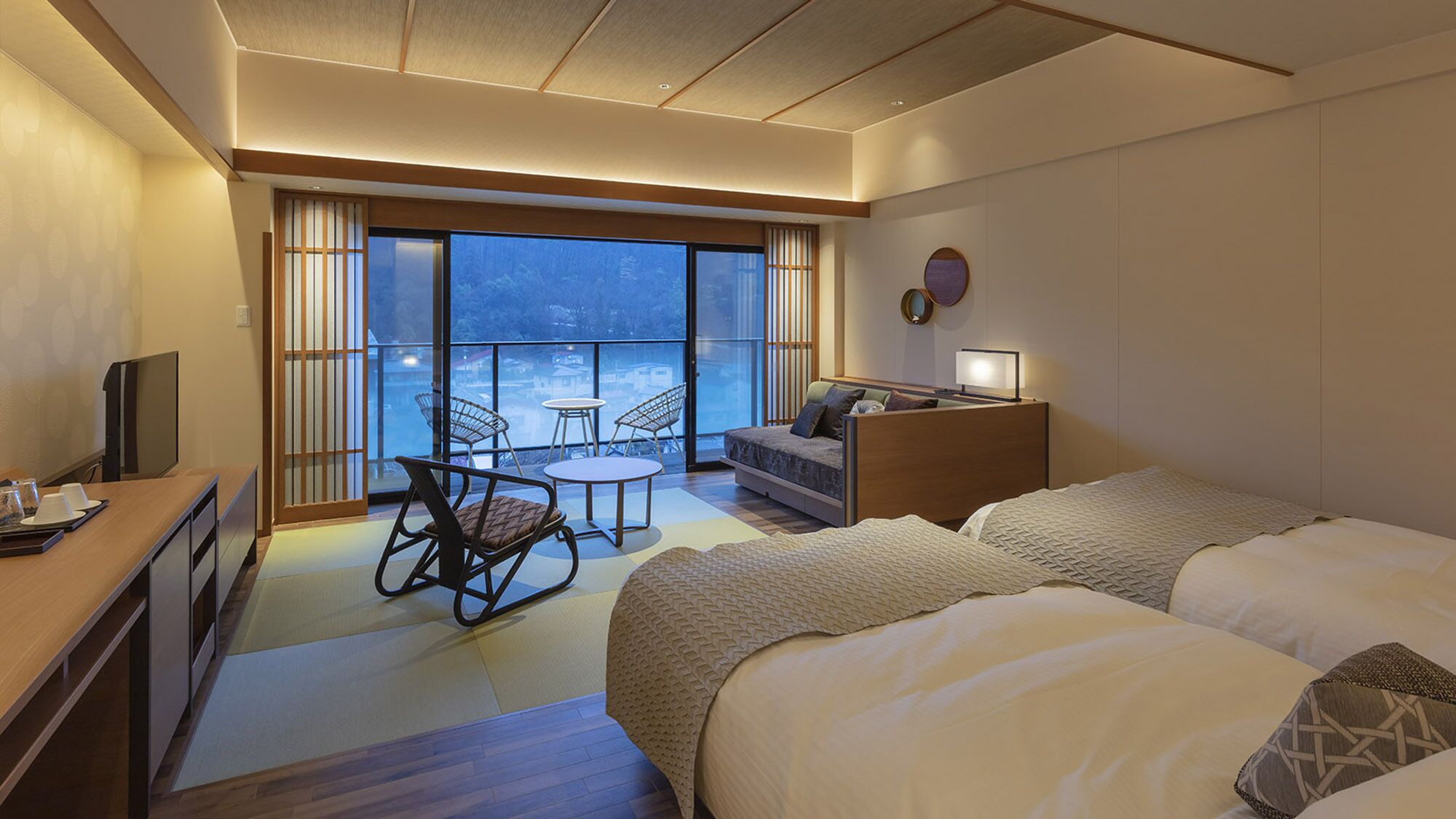 A hotel room with a bed, a desk, a chair, a small table, and a view through large windows.