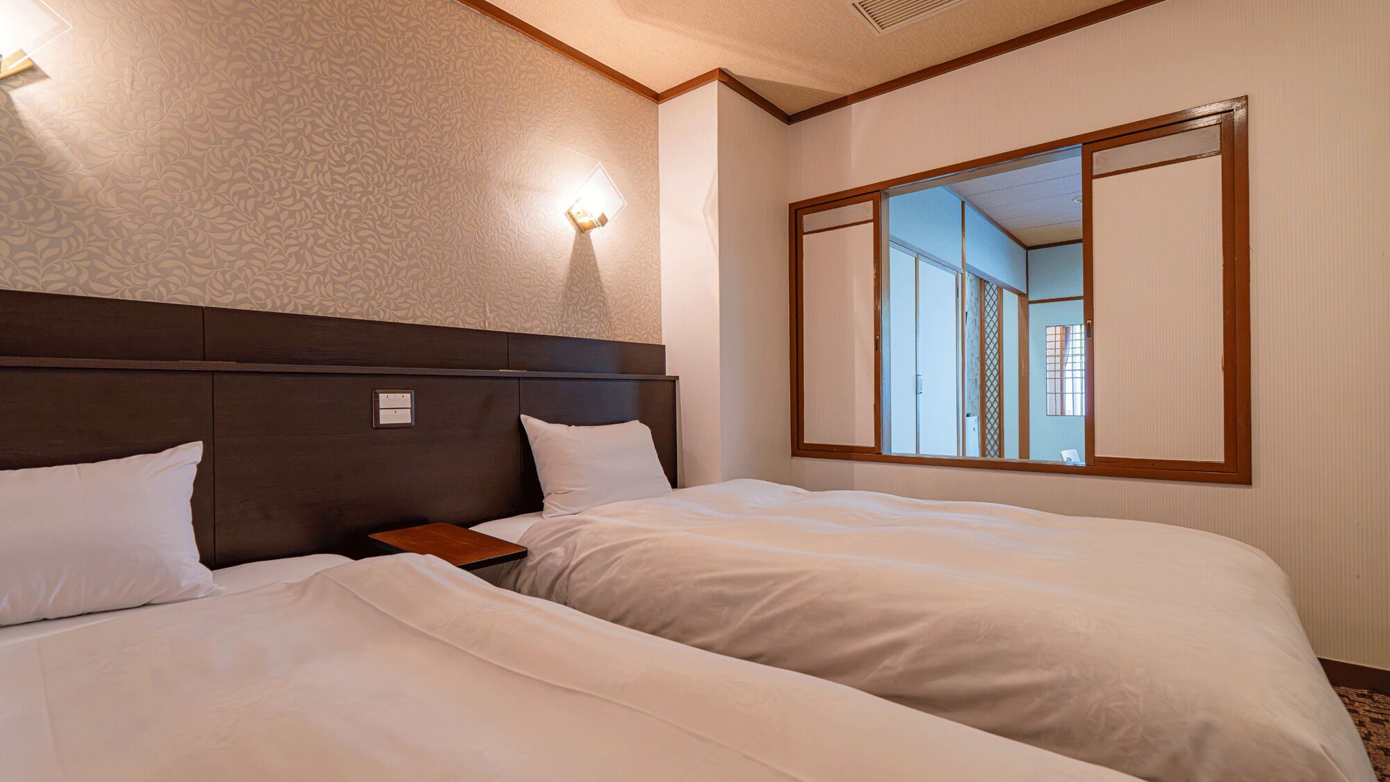 A hotel room with a large bed, a nightstand, a wall-mounted light, and a large mirror reflecting the room.