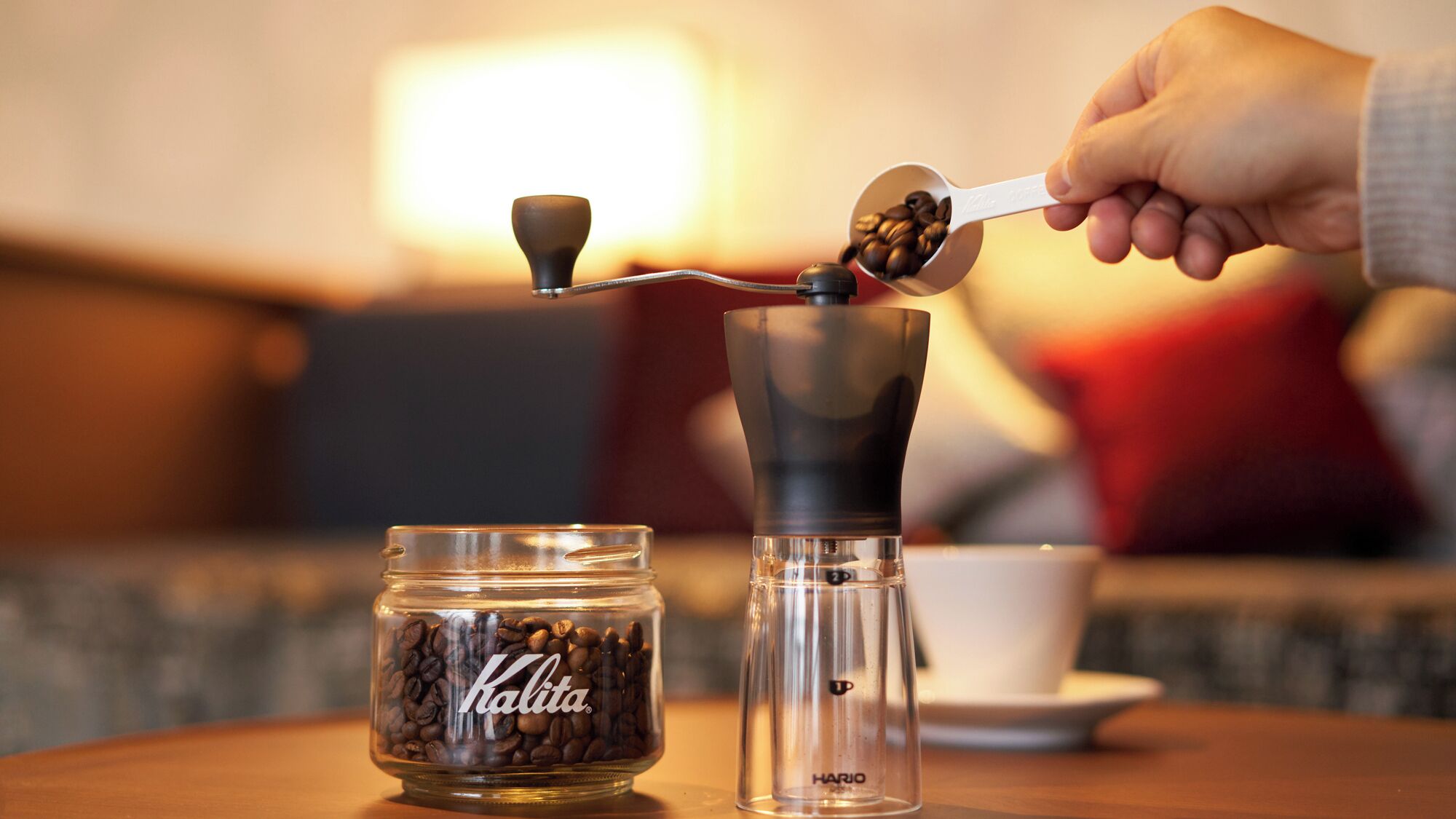 A hand using a coffee grinder to prepare coffee, with a jar of Kalita coffee beans and a clear coffee grinder on a table.