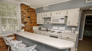 A kitchen with a brick accent wall, white cabinetry, a large island with bar stools, a microwave, stove, and a coffee maker. - At Last Apartment 5 (Murrells Inlet)