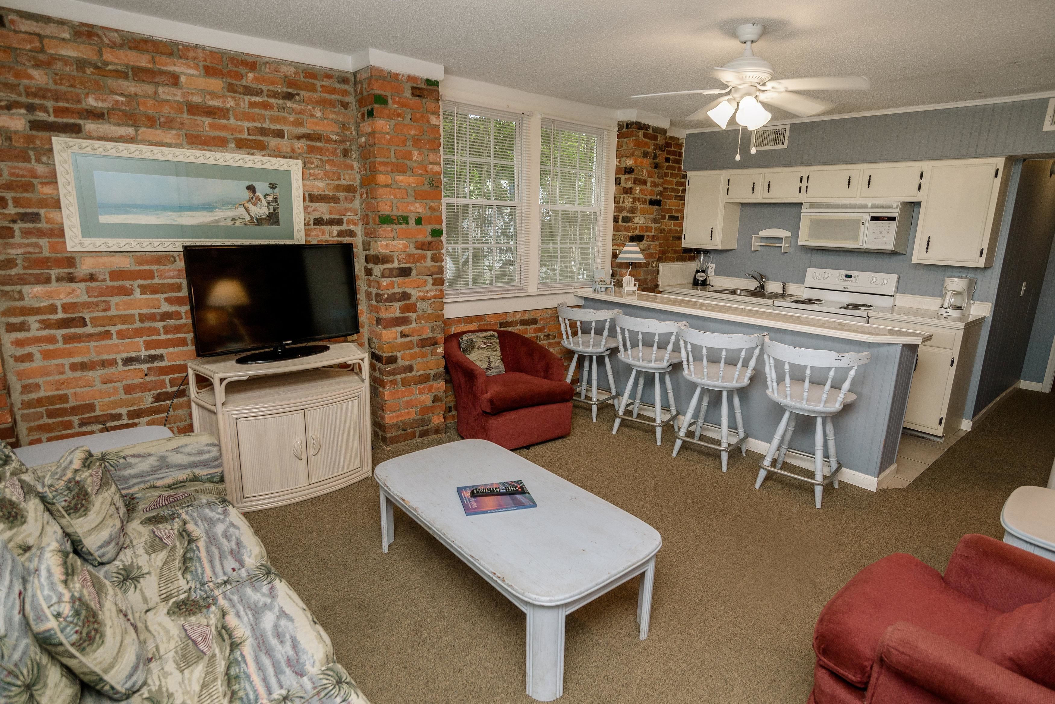 A compact living space with a brick wall, a television, a sofa, a small dining area with bar stools, and a kitchenette.