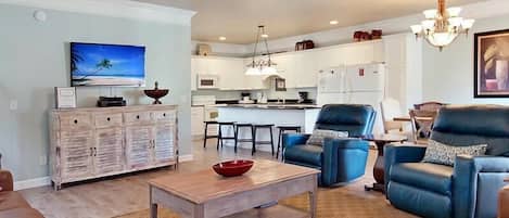 A modern living room with a wooden coffee table, blue armchairs, and a kitchen in the background.