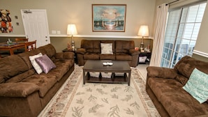 A living room with two brown sofas, a coffee table, and a patterned rug. There are two lamps on side tables, a painting on the wall, and a door in the background.