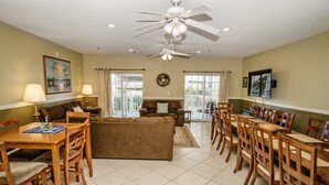 A spacious living room with a brown sofa, wooden dining table, and chairs, a ceiling fan, and a TV mounted on the wall.