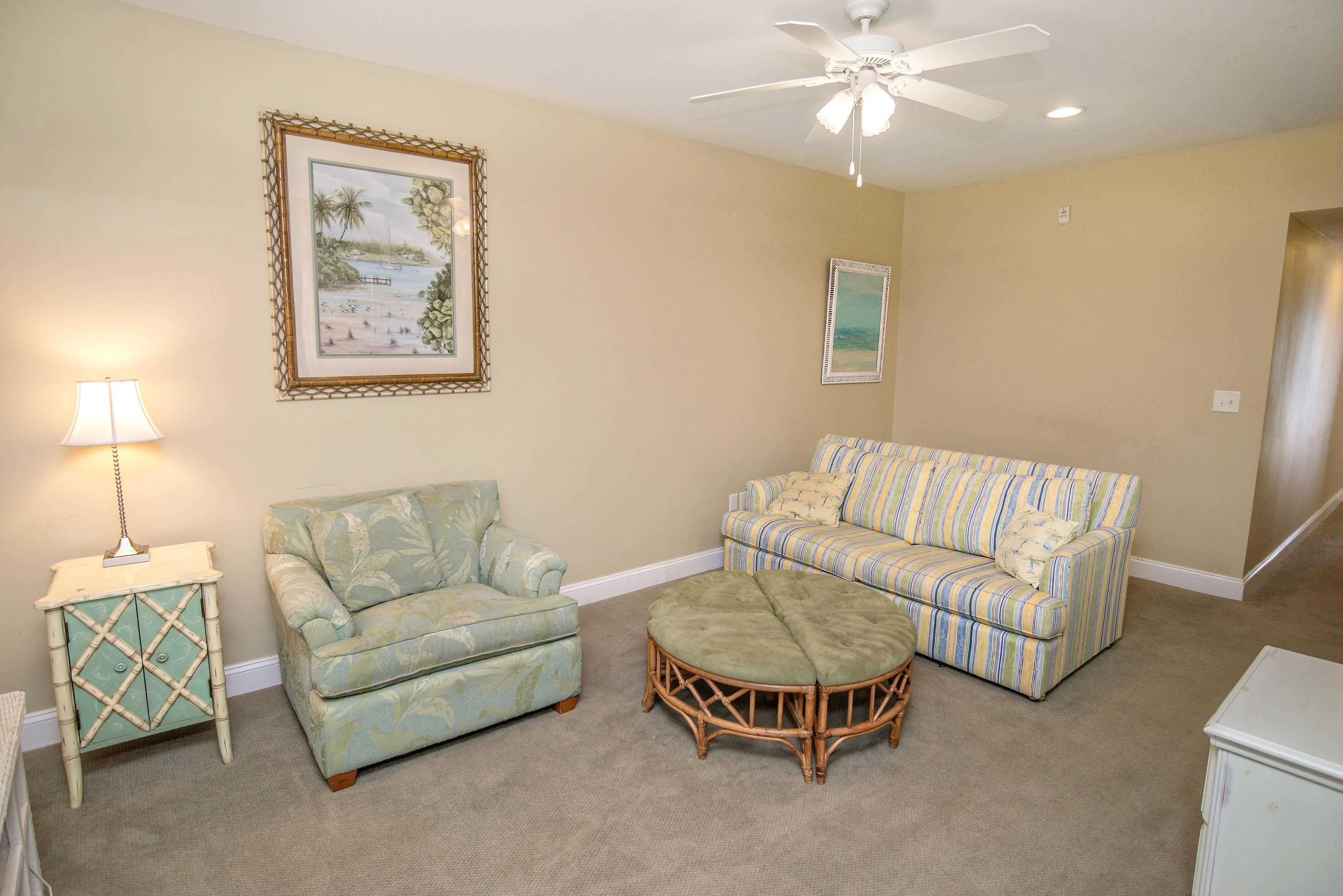 A living room with a green patterned sofa, a striped sofa, a small table with a lamp, and a wicker ottoman.