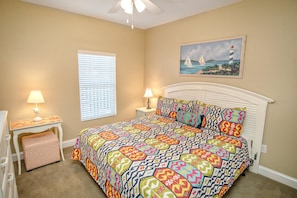 A bedroom with a patterned bedspread, a nightstand with a lamp, a window with blinds, and a painting of a lighthouse on the wall.
