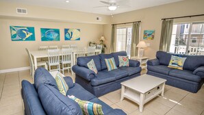 A living room with blue sofas, a white coffee table, and a dining table with chairs. There are paintings on the walls and a window with curtains. - Coconut Grove 204 Villa Villa 5 (North Myrtle Beach)