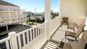 A balcony with white railings, a seating area with two chairs and a table, and a view of neighboring buildings and a street. - Coconut Grove 204 Villa Villa 5 (North Myrtle Beach)