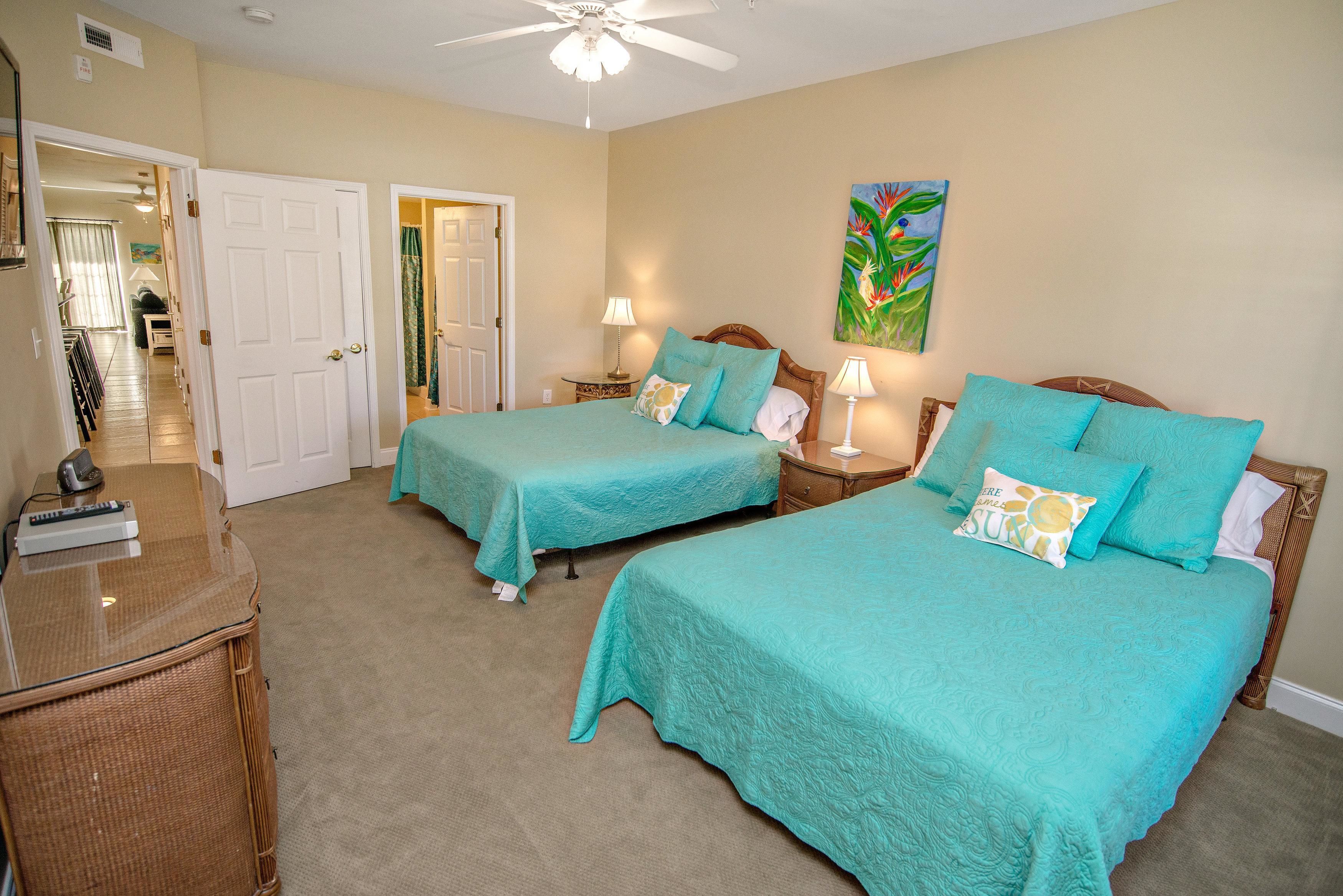 A hotel room with two beds, a ceiling fan, a painting on the wall, and a view of the hallway.