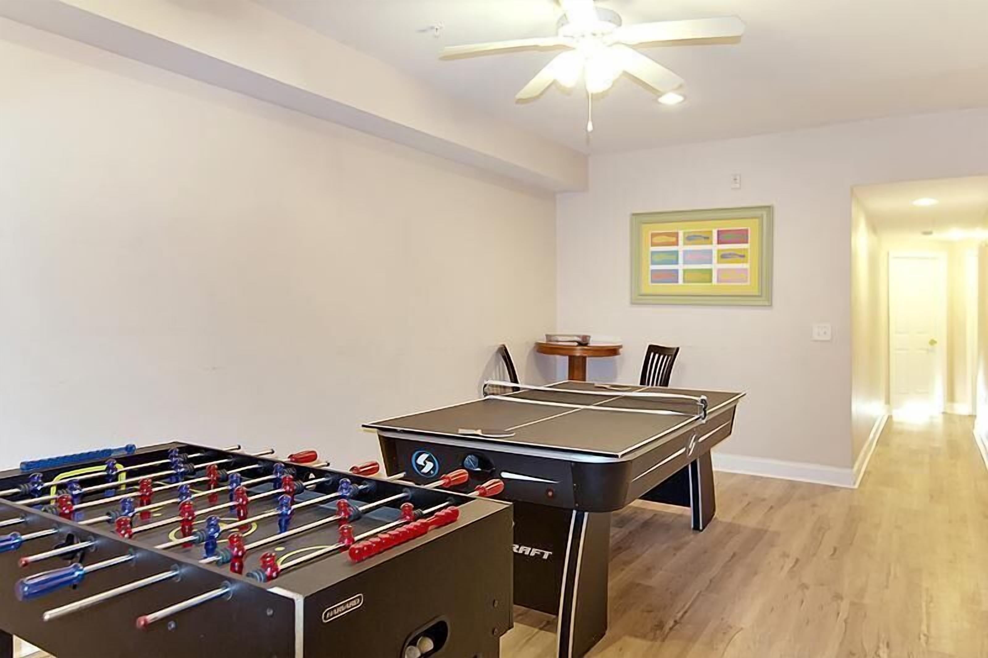 A room with a foosball table, ping pong table, and a ceiling fan.