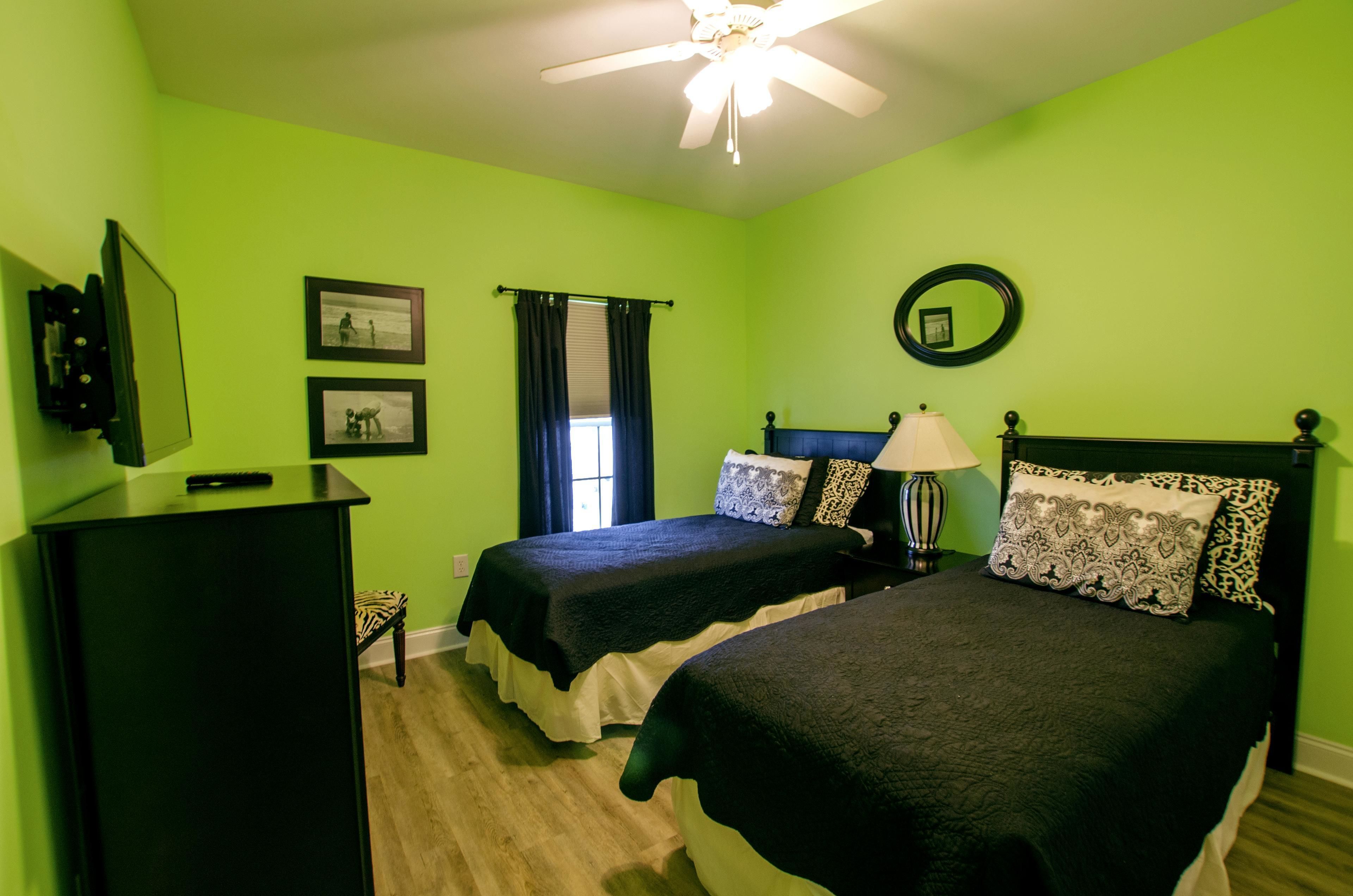 A room with two beds, a ceiling fan, a desk with a chair, and a window with curtains.