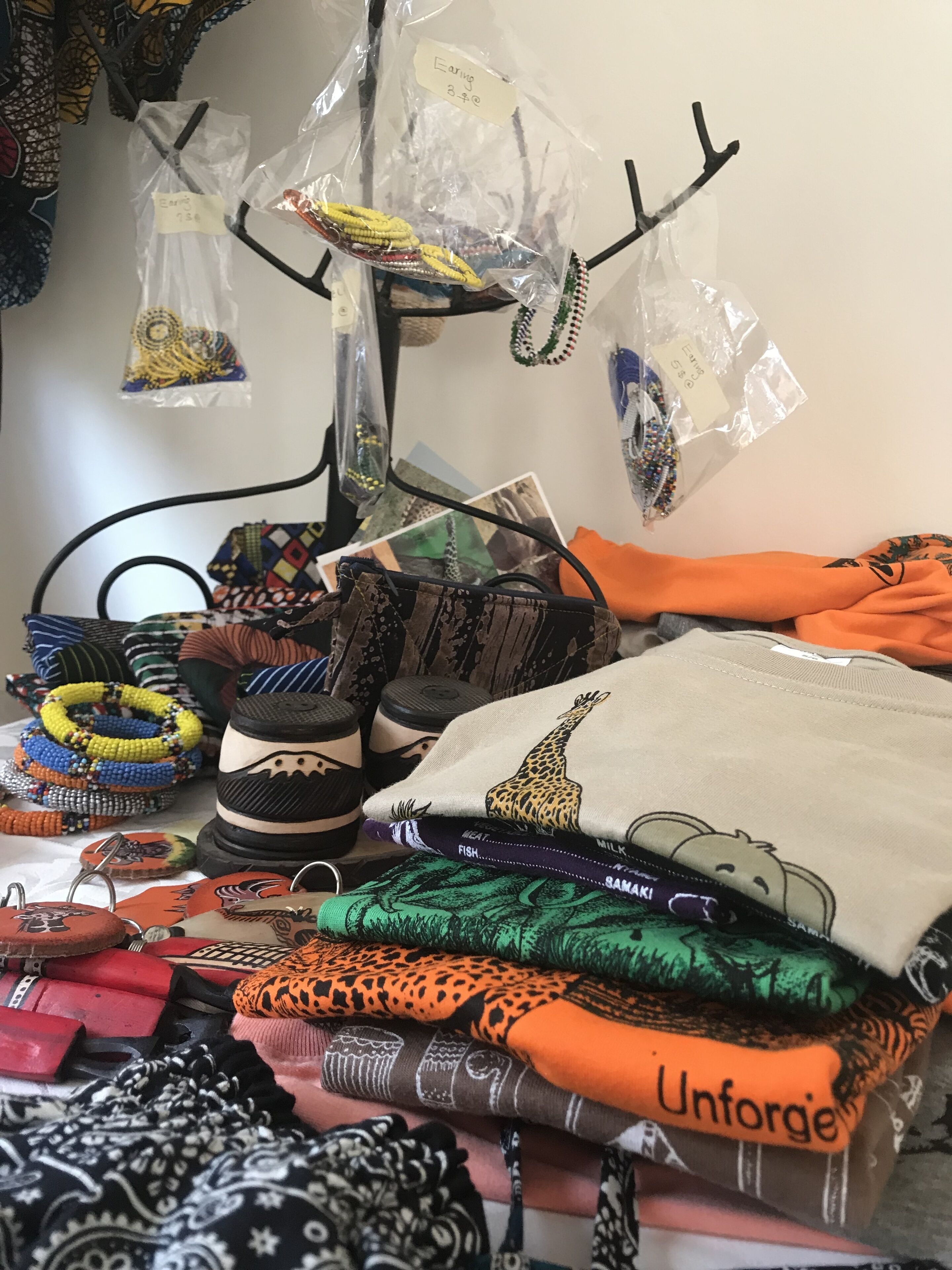 A variety of colorful textiles and accessories, including patterned fabrics, a giraffe-themed pillow, and beaded jewelry, displayed on a table.