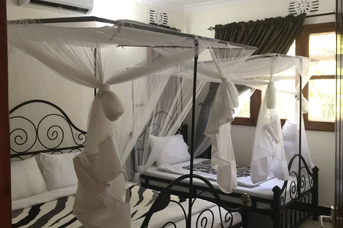 Ein Zimmer mit zwei Betten, jeweils mit Baldachin. An der Wand befindet sich eine Klimaanlage, ein Fenster mit Vorhängen und eine Tür mit Schlüsselanhänger.