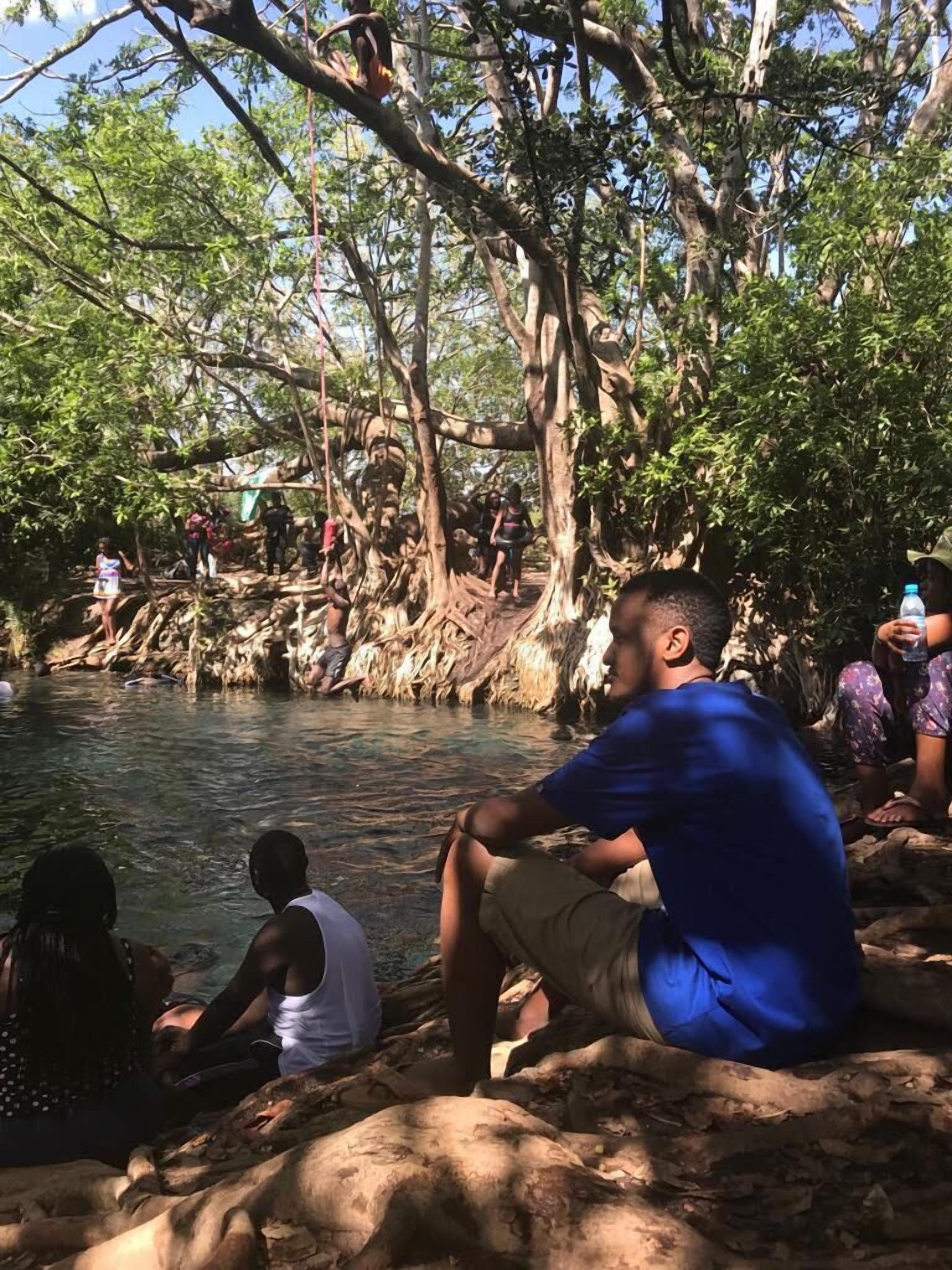 A group of people sitting by a river surrounded by trees.