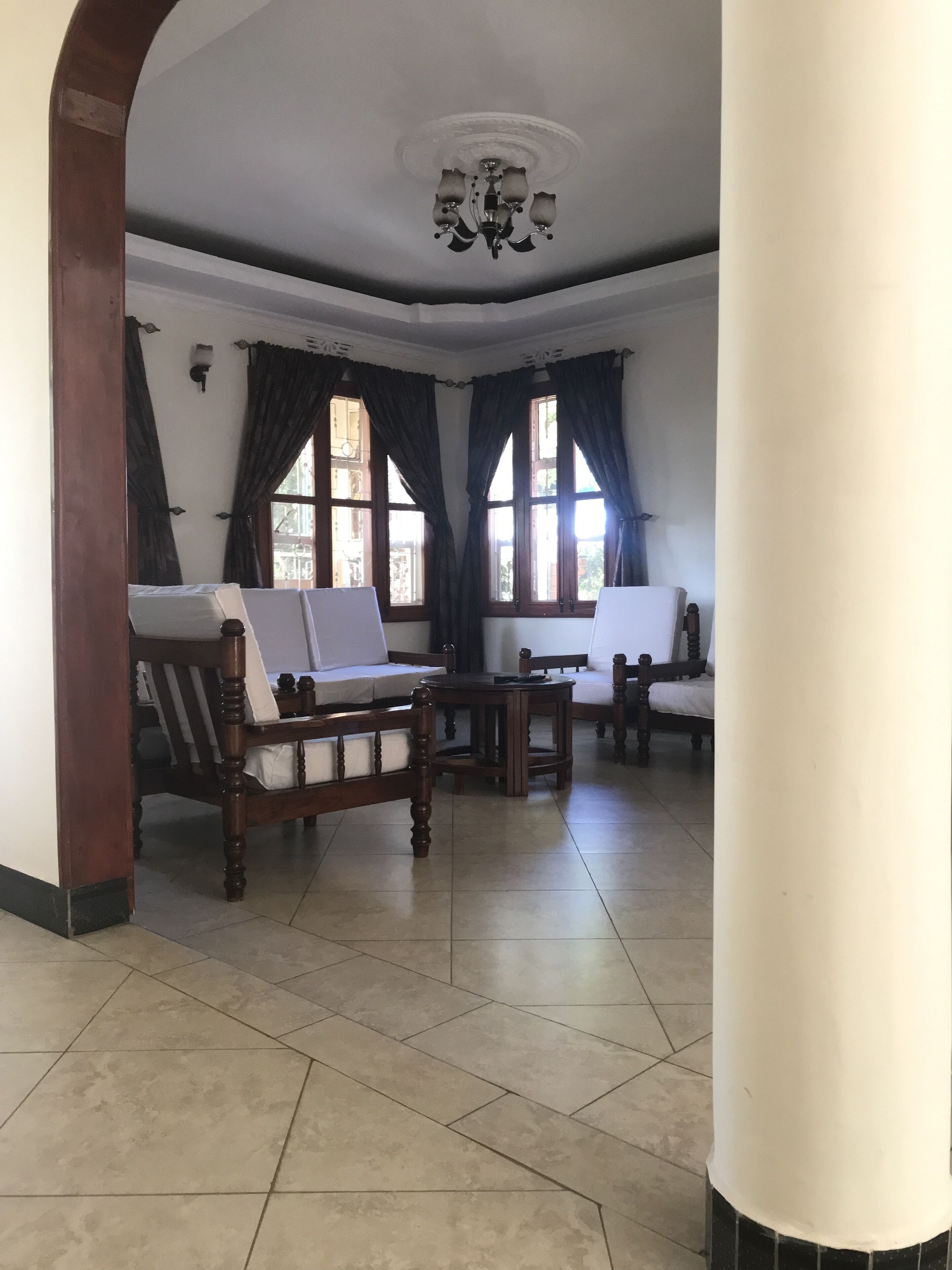 A spacious room with a high ceiling, a chandelier, large windows with curtains, and a tiled floor.