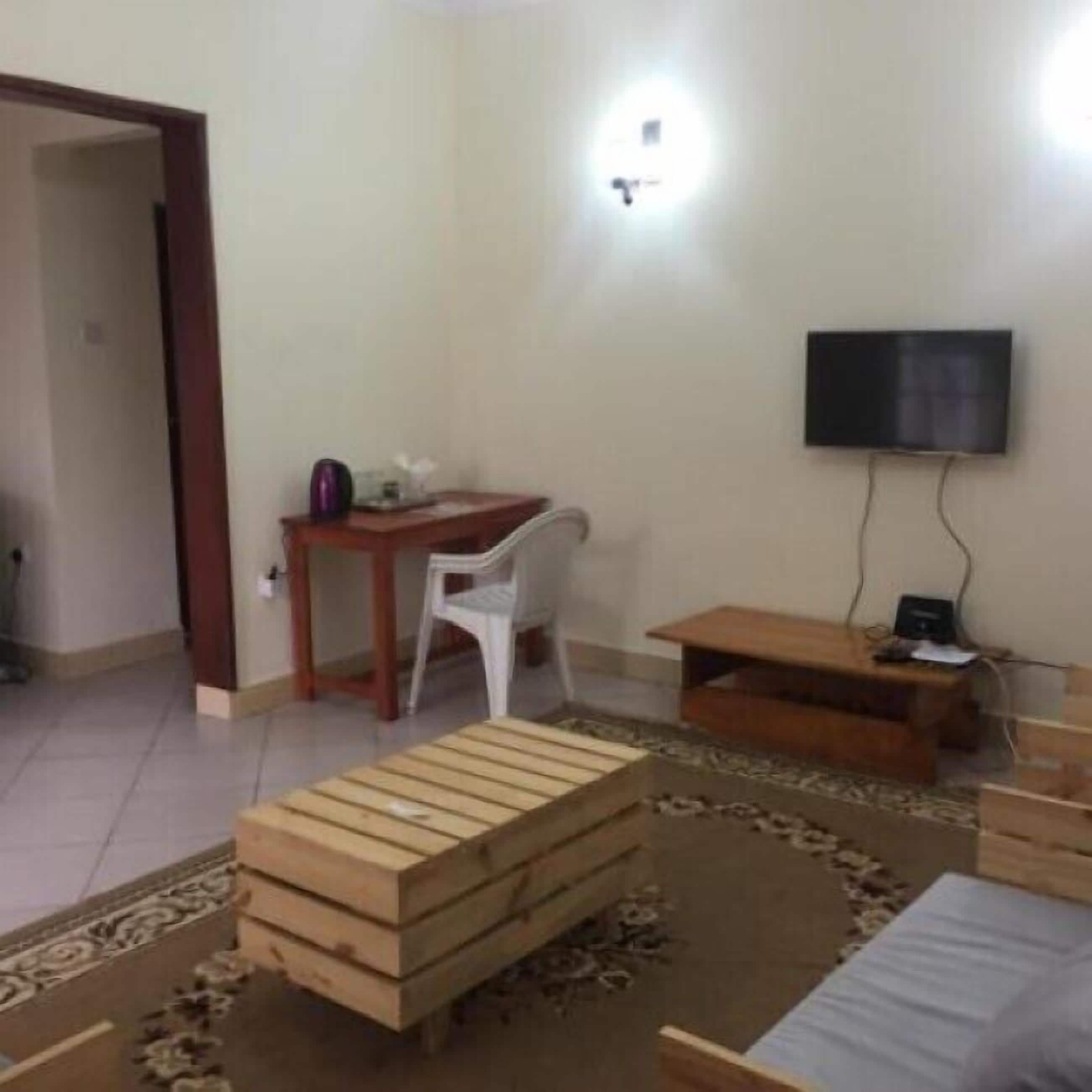 A room with a wooden bench, a small table, a white chair, a TV mounted on the wall, and a door leading to another room.