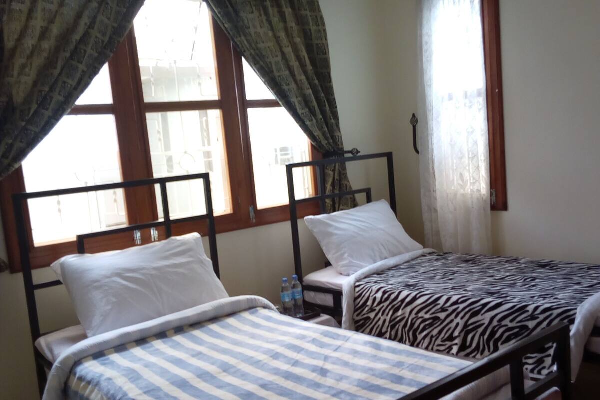 Ein Zimmer mit zwei Betten, jedes mit einer anderen Bettdecke. Es gibt Fenster mit Vorhängen und einen Heizkörper an der Wand.