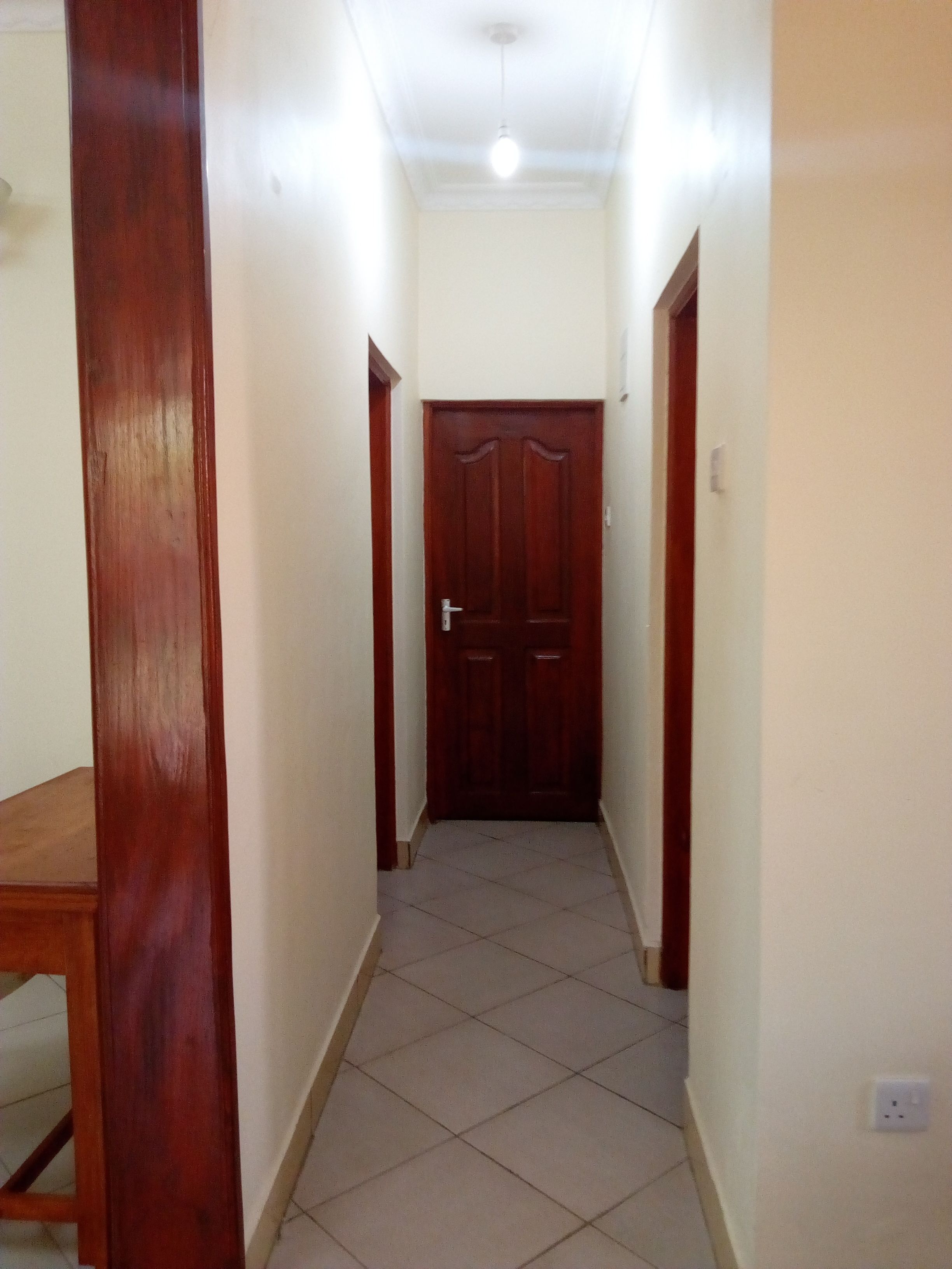 A narrow hallway with a wooden door at the end, tiled floor, and white walls.