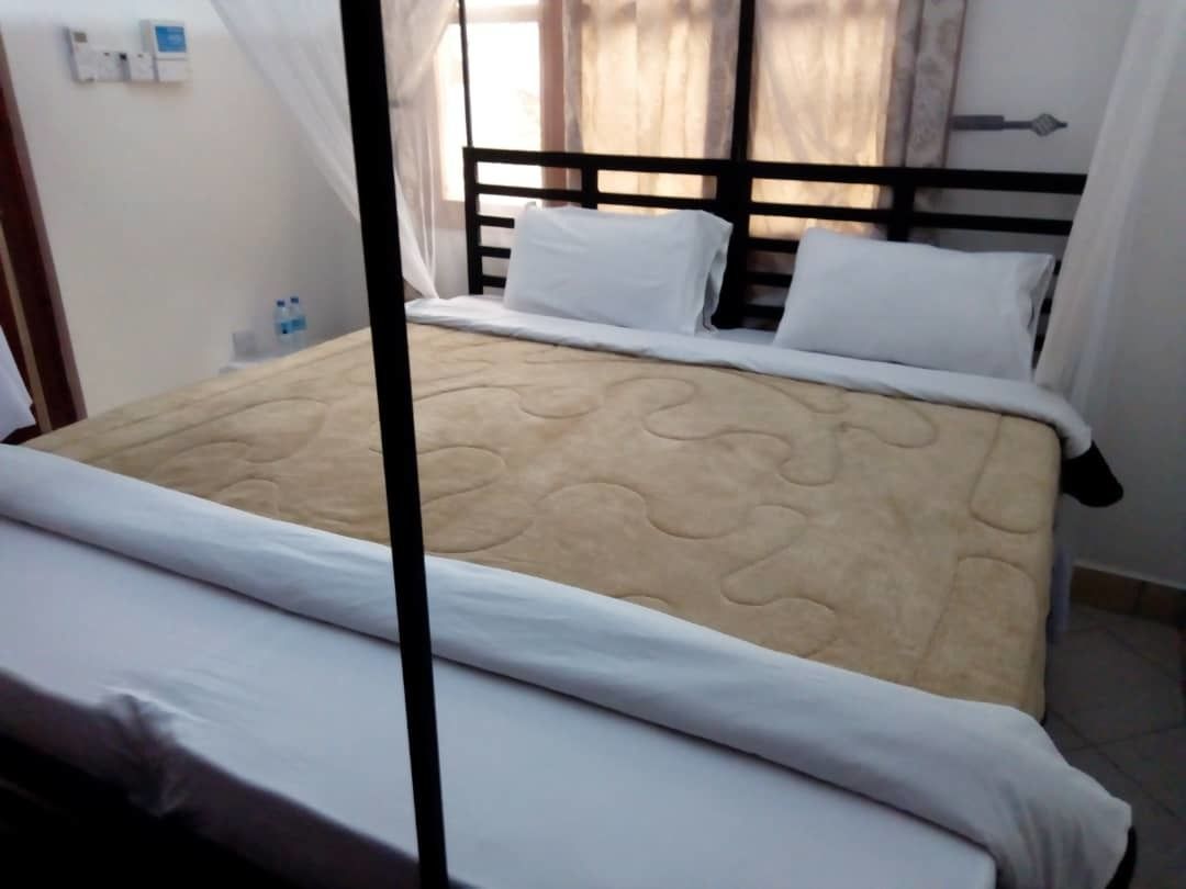 A neatly made bed with white bedding and a beige quilt, a black headboard, and a bedside table with a water bottle.