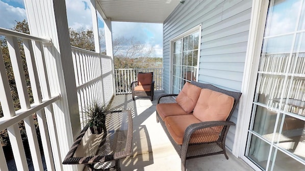 A covered porch with a wicker chair and a cushioned bench, a potted plant, and a white railing.