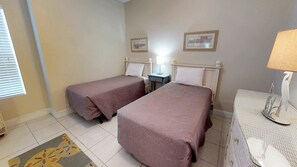 A hotel room with two single beds, a small bedside table with a lamp, a wall-mounted picture frame, and a patterned rug.
