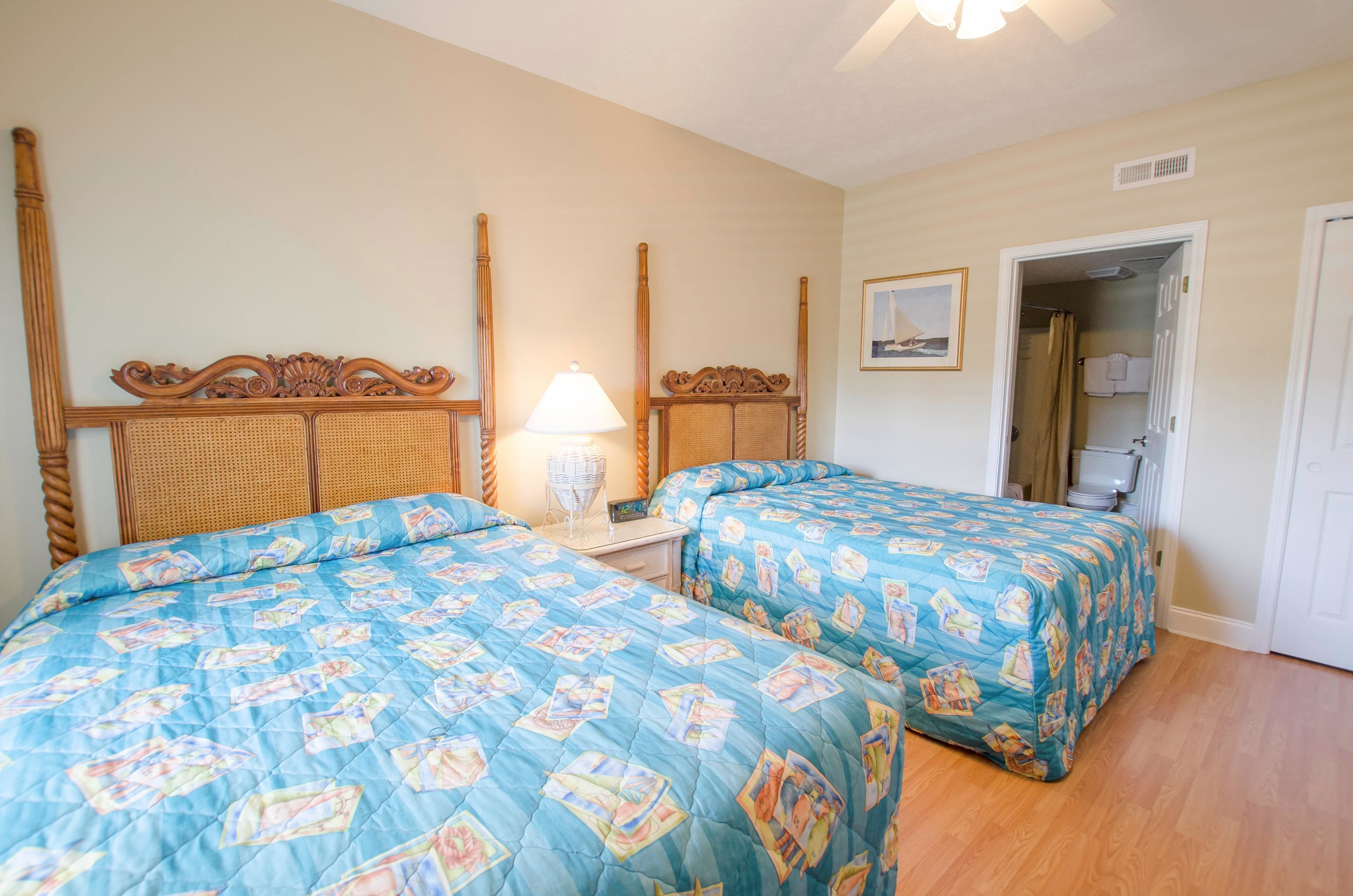 A room with two beds, a nightstand, a lamp, a framed picture, and a door leading to a bathroom.