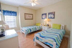 A bedroom with two single beds, a desk, a chair, a ceiling fan, and artwork on the walls.
