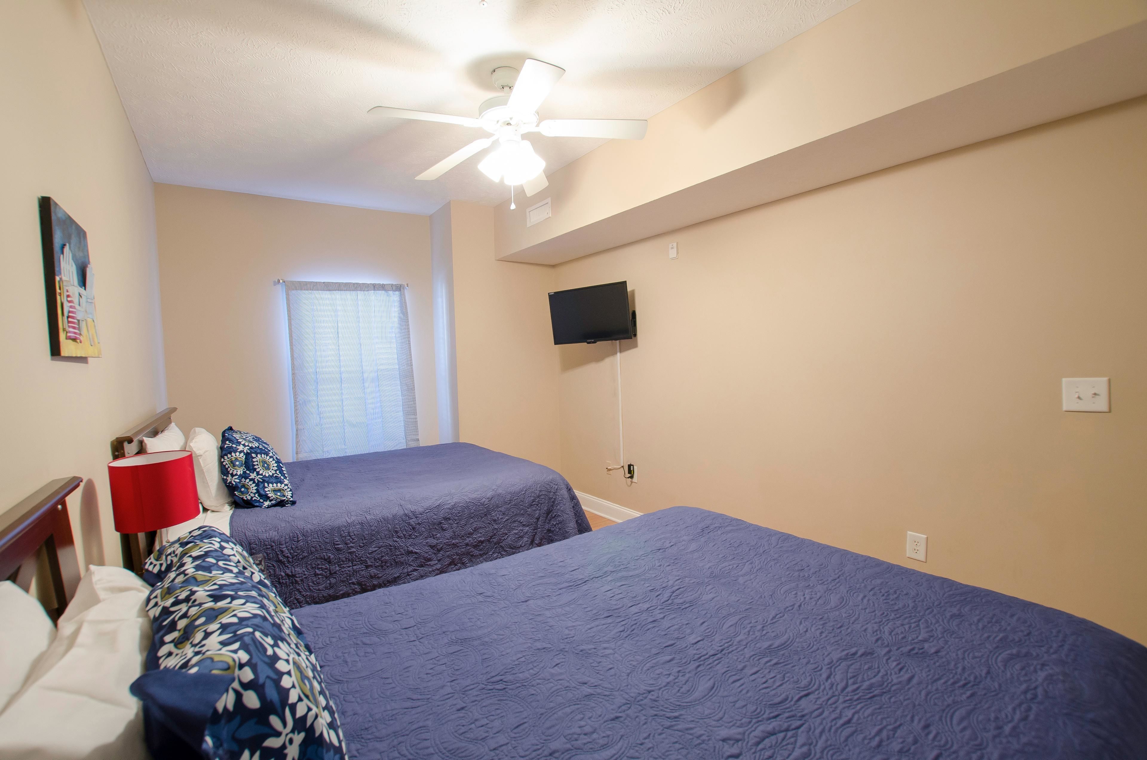 A hotel room with two beds, a ceiling fan, a TV mounted on the wall, and a window with curtains.