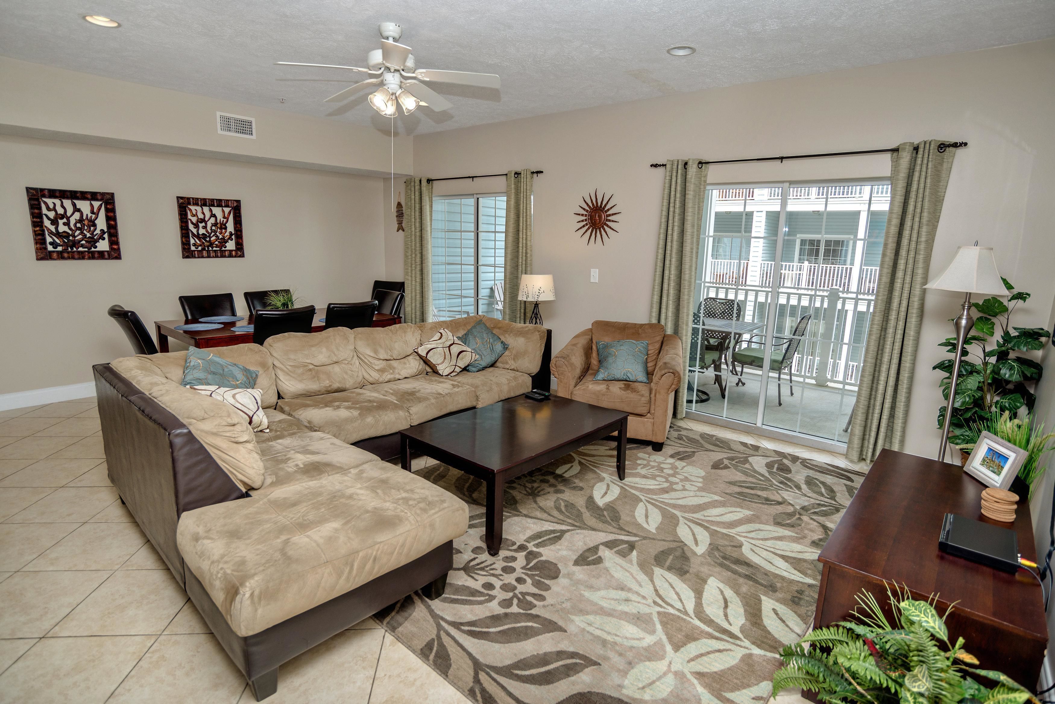 A living room with a sectional sofa, a coffee table, and a dining area with chairs. There is a balcony with a railing and a view of the outside.