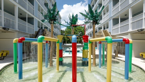 Un área de juegos acuáticos con estructuras coloridas y una piscina, rodeada por un edificio de varios pisos con balcones.