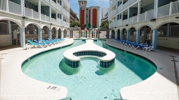 Un espace piscine d’hôtel avec une fontaine centrale, de nombreux fauteuils de relaxation et, au fond, un beffroi.