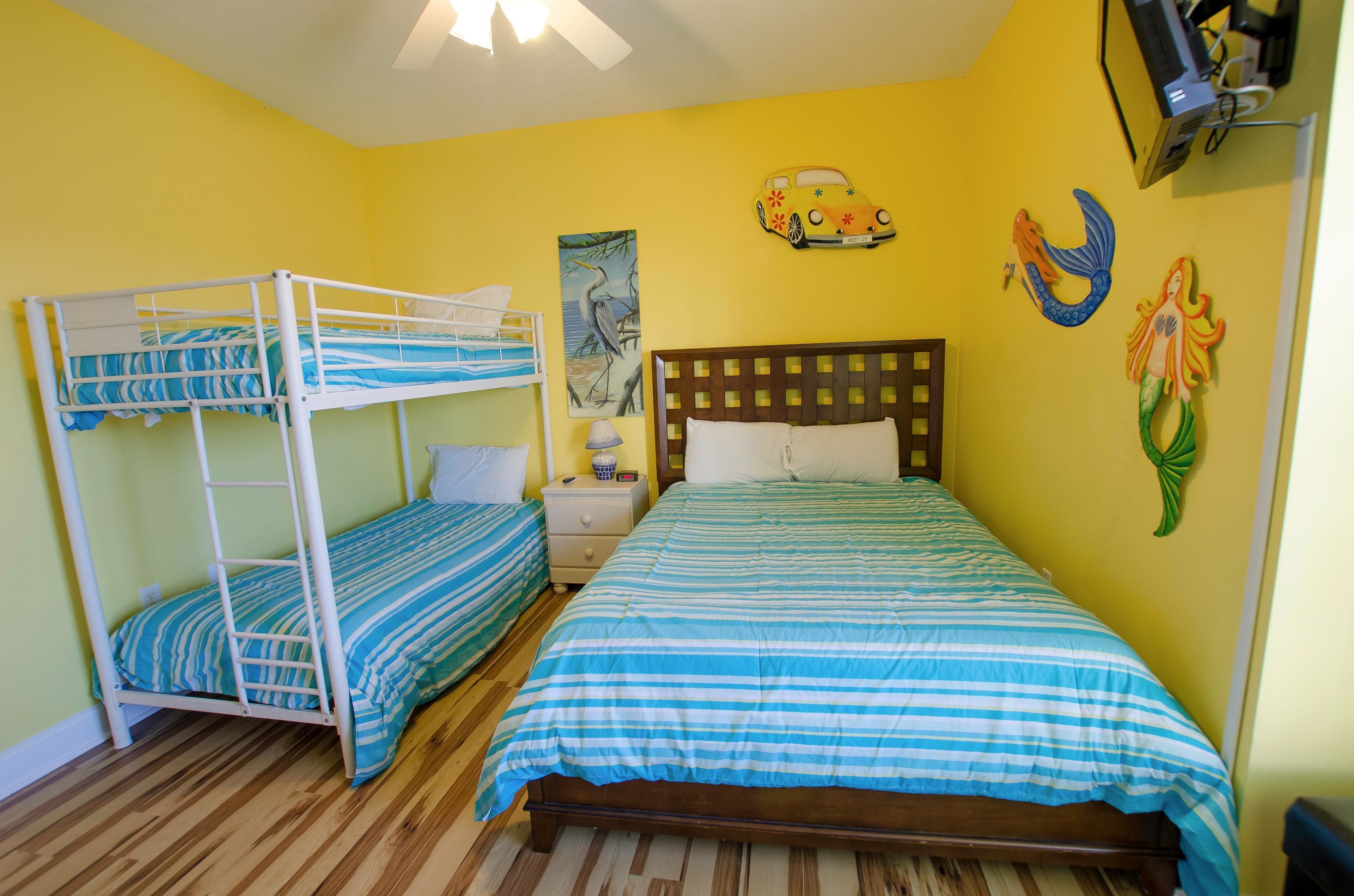 A room with two bunk beds, a wooden bed, a nightstand, a lamp, and wall art including a car and mermaid.