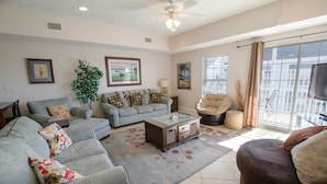 A spacious living room with a large sofa, a coffee table, and a TV. There is a plant and artwork on the walls, and a balcony with a view. - Myrtle Beach 401A Villa Villa 4 (Myrtle Beach)
