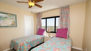 A room with two beds, each with a floral bedspread, a ceiling fan, a window with curtains, and a painting on the wall. - The Palms 1701 Villa Villa 4 (Myrtle Beach)