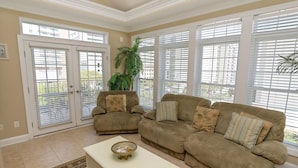 A living room with beige walls, a beige sofa, a white coffee table, and a large window with blinds.