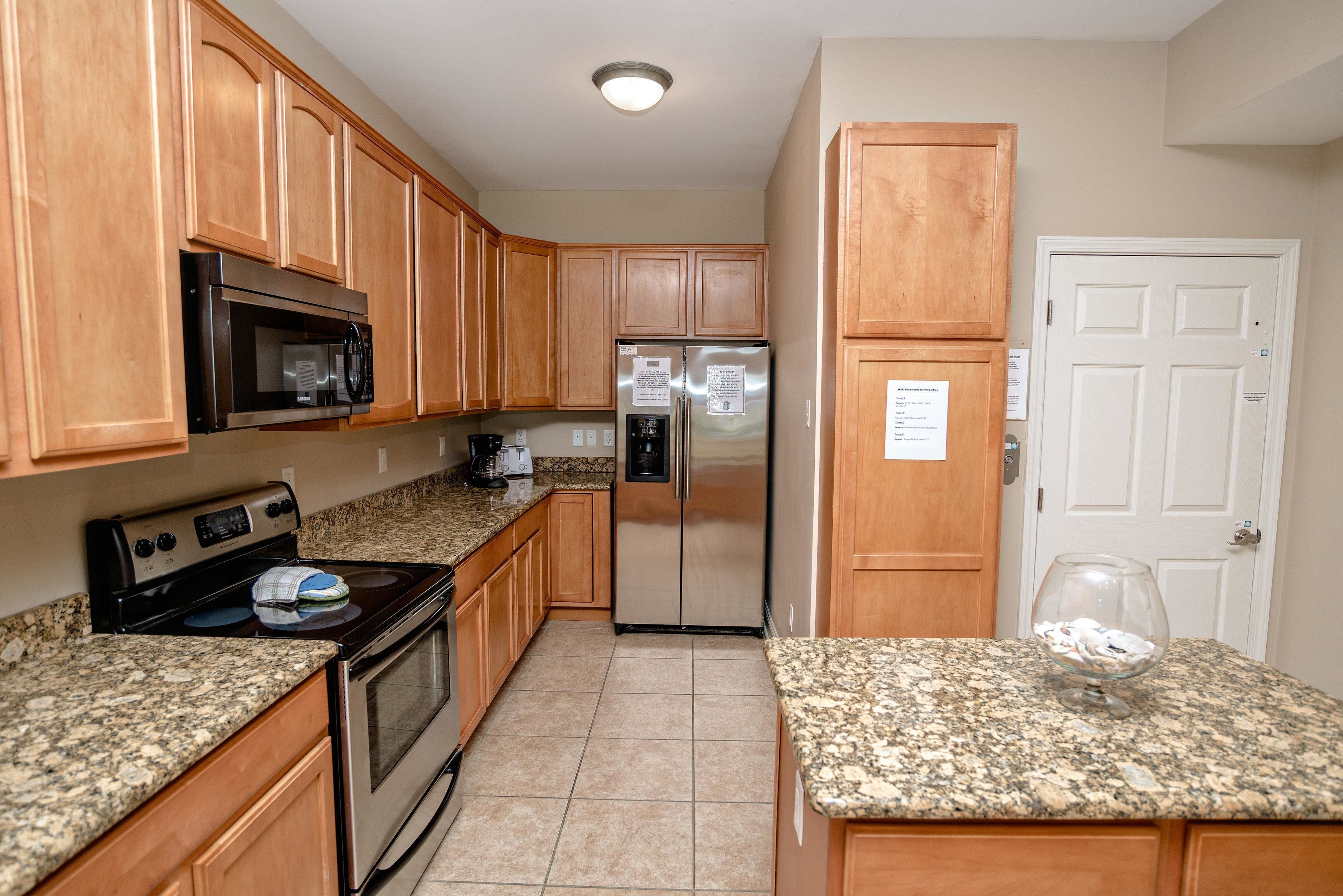 A kitchen with wooden cabinets, a stainless steel refrigerator, a microwave, an oven, granite countertops, and a door with a sign.
