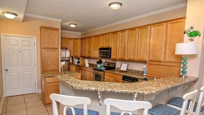 A kitchen with wooden cabinets, granite countertops, a microwave, a stove, and a sink. There are four bar stools with blue cushions. - Tuscan Ocean Vista D Holiday Home Holiday home 8 (Myrtle Beach)