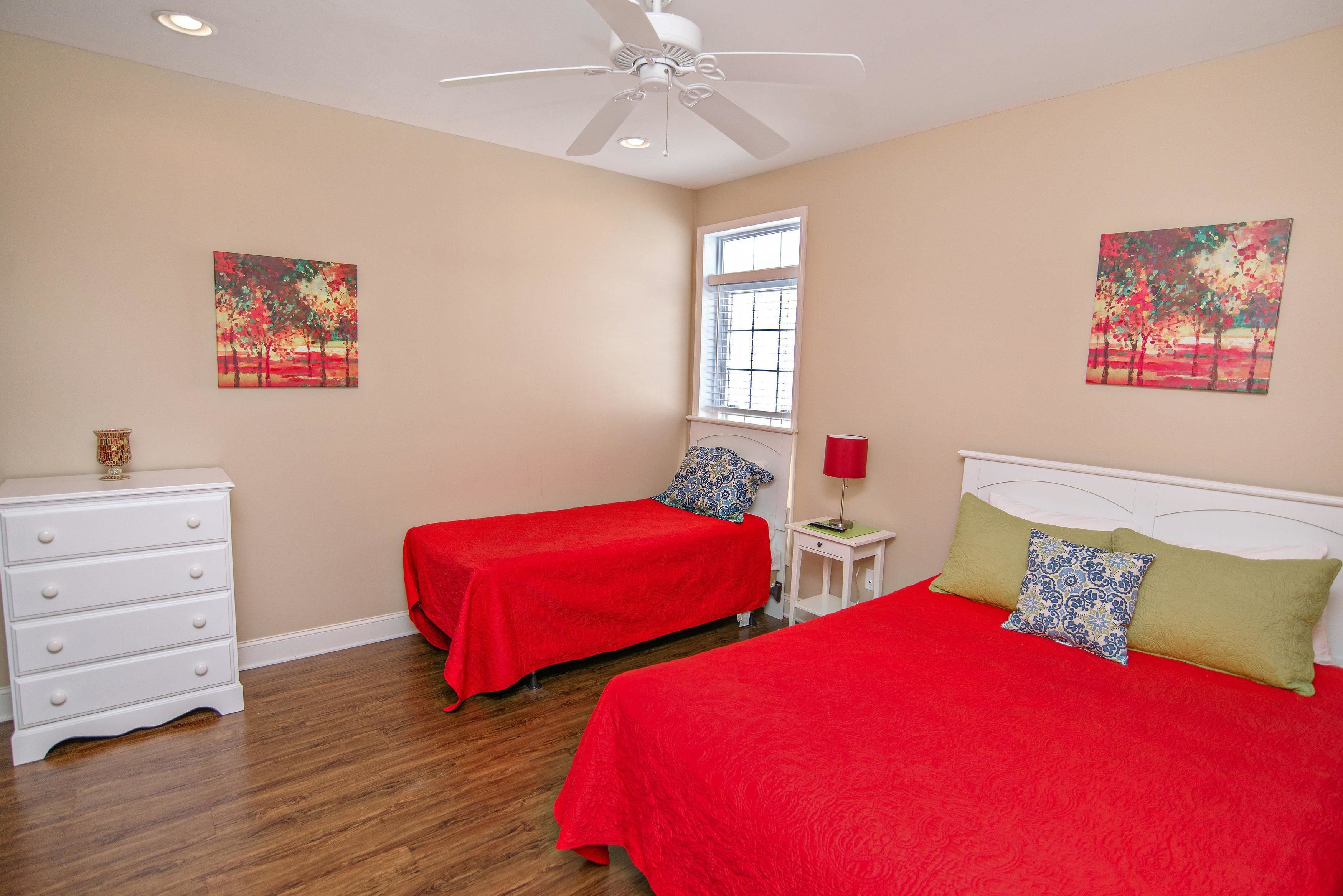 A bedroom with two beds, a dresser, a ceiling fan, and two paintings on the walls.