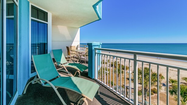 A balcony with two green lounge chairs and a table, overlooking a beach with palm trees and a clear blue sky.