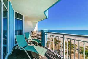A balcony with two green lounge chairs and a table, overlooking a beach with palm trees and a clear blue sky.