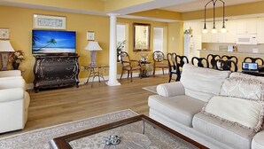 A living room with a sofa, a coffee table, and a TV. There is a dining area with chairs and a table. The kitchen is visible in the background.