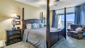 A four-poster bed with a dark finish, a bedside table with a lamp, and a chair with a cushion.