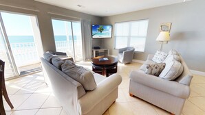 A spacious living room with a large TV, two sofas, a coffee table, and a view of the beach.