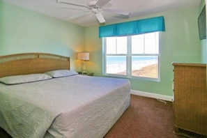 Ein Schlafzimmer mit Bett, Deckenventilator, Fenster mit Blick auf den Strand und Nachttisch mit Lampe.