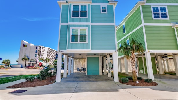 A two-story building with a green and blue facade, numbered 2701 and 2709, featuring a covered entrance with a palm tree and small shrubs. - South Beach Cottages - 2701 Apartment 4 (Myrtle Beach)