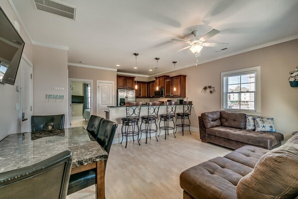 A spacious living area with a dining table, bar stools, a brown leather sofa, and a ceiling fan.